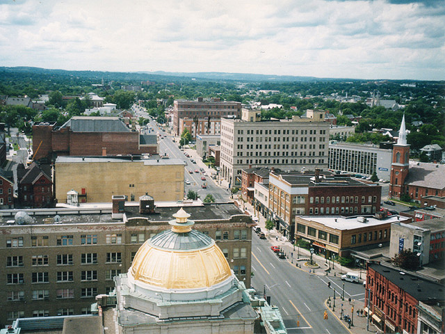 Aerial view above Genesee Street showing historic downtown Utica with buildings, storefronts, and traffic along the main street.